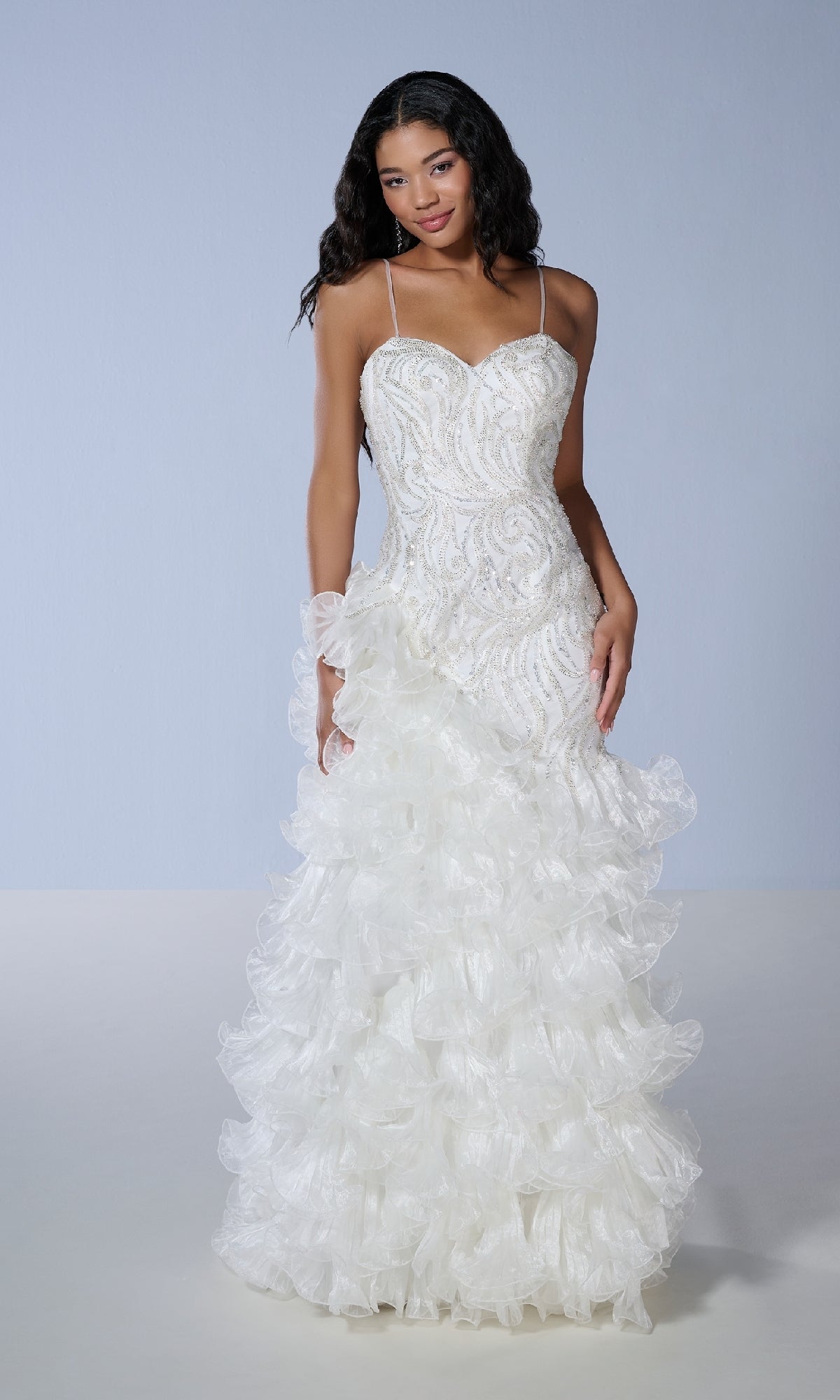 A woman stands against a plain light background wearing the Tiffany 16234, a long white beaded tulle prom dress with spaghetti straps and a ruffled mermaid skirt.